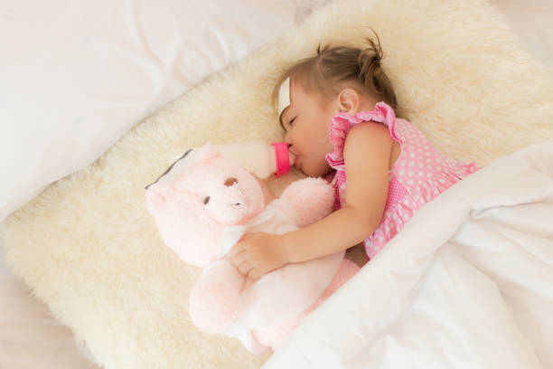 istock Baby drinking water or milk from a small bottle and lying on bed while hugging pink teddy bear in the bedroom because the baby having fever and attach cooling gel pad on her forehead for relief fever. 898298964