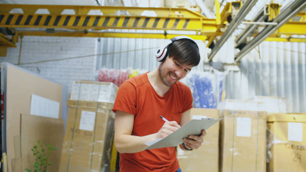 giovane lavoratore felice nel magazzino industriale ascoltando musica e ballando durante il lavoro. l'uomo in cuffia si diverte sul posto di lavoro. - clipboard men sales occupation casual foto e immagini stock