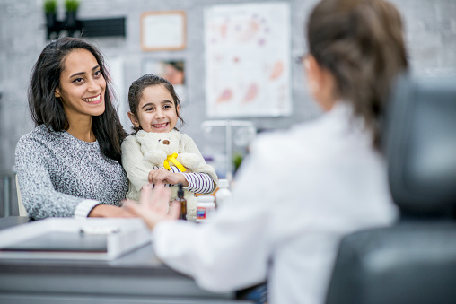 A young girl and her mother are in a doctor's office. The daughter is holding a stuffed teddy bear. They are happily talking to their doctor.