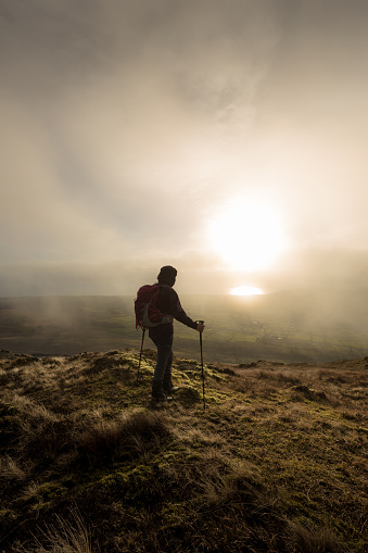 single full body hill walker standing on grassy mountain looking at the sunset with walking poles and a misty moody sky