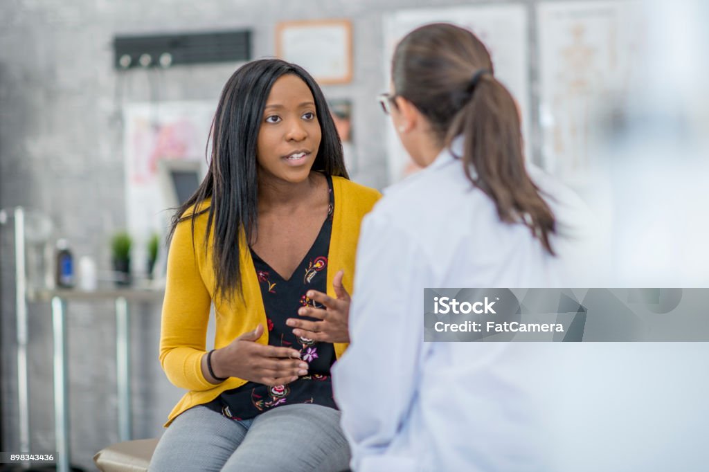 Patient Describing Symptoms A woman of African descent and her doctor are indoors in a medical clinic. The woman is sitting and describing her symptoms to the doctor. Doctor Stock Photo