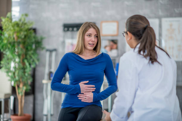 Woman With A Stomachache A Caucasian woman and her doctor are indoors in a medical clinic. The woman is holding her stomach and describing her stomachache symptoms. stomachache stock pictures, royalty-free photos & images