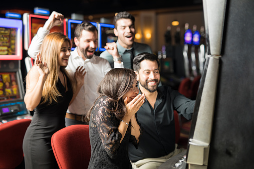 Beautiful Hispanic woman looking excited about hitting the jackpot in a slot machine while her friends celebrate with her