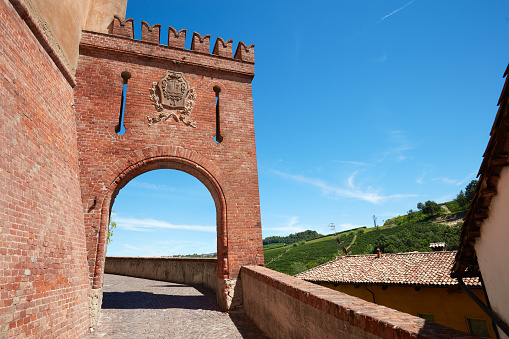 Barolo, Italy - August 6, 2016: Barolo medieval castle entrance arch in red bricks and emblem with empty street in a sunny summer day, blue sky in Barolo, Italy. The Langhe area in Piedmont is Unesco World Heritage Site.