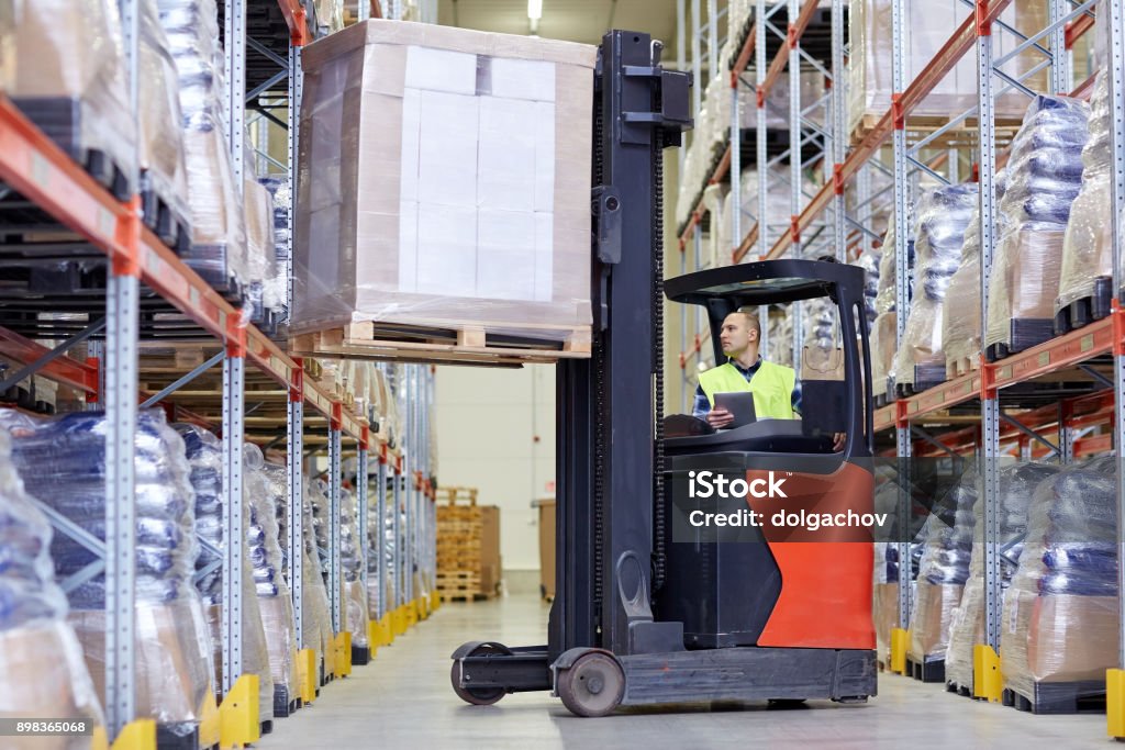 man with tablet pc operating forklift at warehouse wholesale, logistic, loading, shipment and people concept - man or loader with tablet pc computer and forklift or loader loading boxes at warehouse Forklift Stock Photo