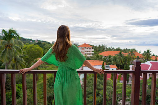 A young woman in green dress is standing on a balcony in tropical country and look forward on sea