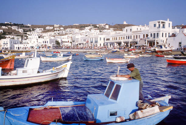 Tourists crowd the waterfront on Mykonos, Greece, as a fisherman maintains equipment on his small fishing boat in the harbor. August 1984. Soft focus, scanned film.