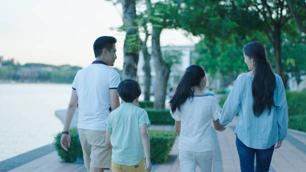 Rear view of Asian family smiling & walking on waterfront promenade at dusk stock photo