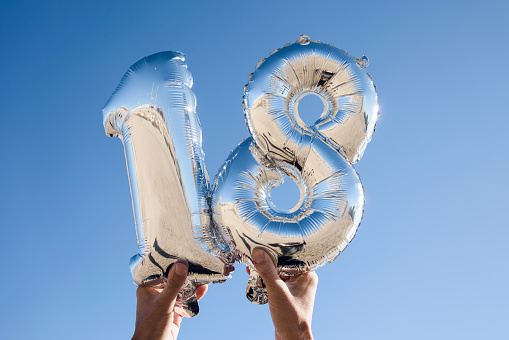 closeup of the hands of a young man holding some silvery number-shaped balloons forming the number 18 against the blue sky