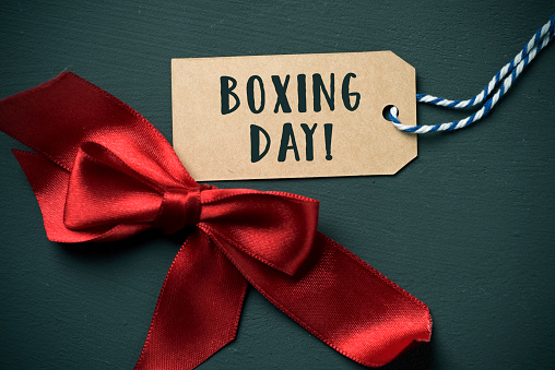 closeup of a red satin ribbon bow and the text boxing day in a brown swing tag, on a dark green background