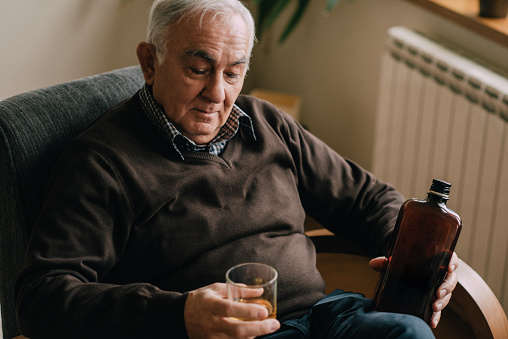 The elderly senior man is seen drinking whiskey in the living room.