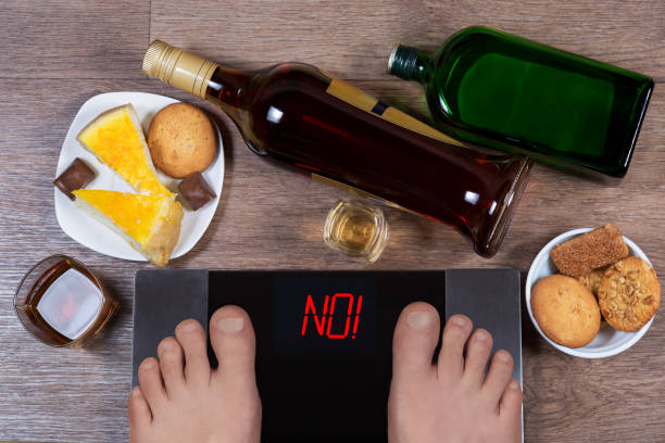Male feet on digital scales with word no on screen. Bottles and glasses of alcohol, plates with sweet food. Concept of consequences of unhealthy lifestile. Morning after party. stock photo