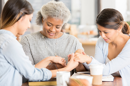 Diverse female mixed age friends hold hands as they pray together. They are meeting in a local cafe for a Bible study.