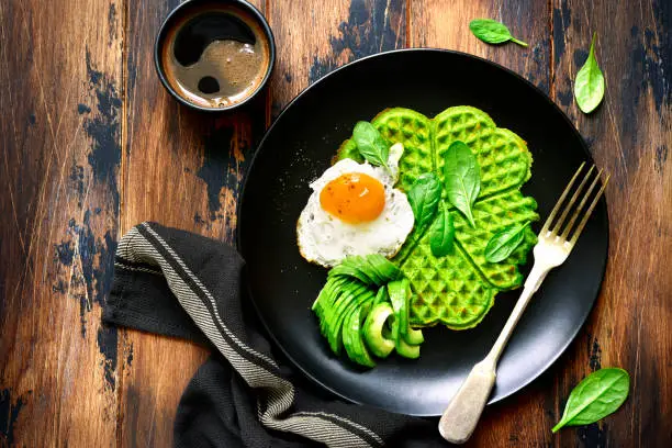 Spinach cheese waffles with fried egg and avocado on a black plate over dark rustic wooden background.Top view.