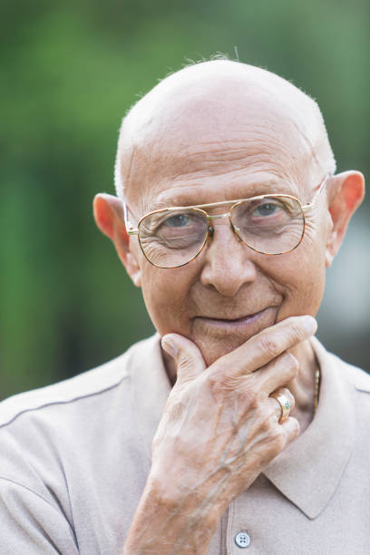 Portrait of an Hispanic man in his 90s. He is standing outdoors, hand on his chin, looking at the camera.