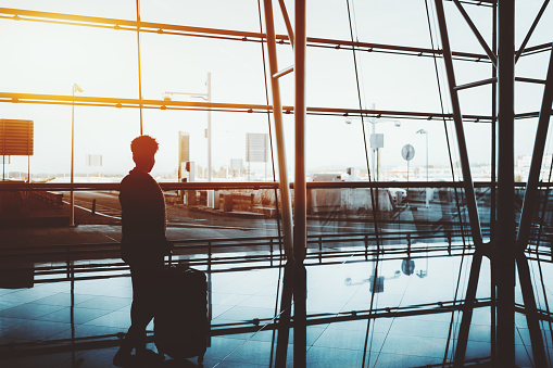 Silhouette of curly black female standing indoors with a huge rolling bag in front of panoramic window and facade of modern airport terminal with strong reflections on the floor and sunny day outside