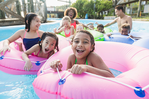 A multi-ethnic group of children, 7 to 11 years old, and two young adults, having fun at summer camp at a water park. They are floating on inner tubes on the lazy river. The focus is on the mixed race African American and Caucasian girl in the foreground.
