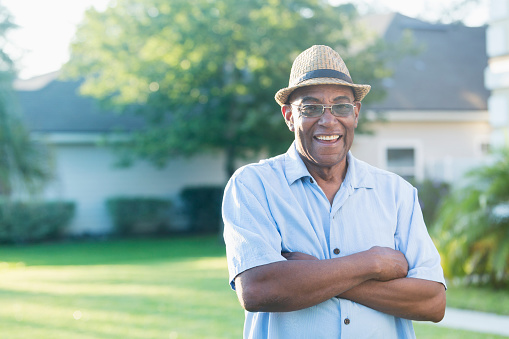 A senior African-American man standing with his arms crossed in his front yard, smiling at the camera. He is relaxed and confident, wearing a stylish hat and eyeglasses.