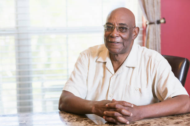 Senior African-American man sitting at table by window A senior African-American man in his 60s sitting at a table by a window with hands clasped, looking at the camera through eyeglasses. 65 69 years stock pictures, royalty-free photos & images