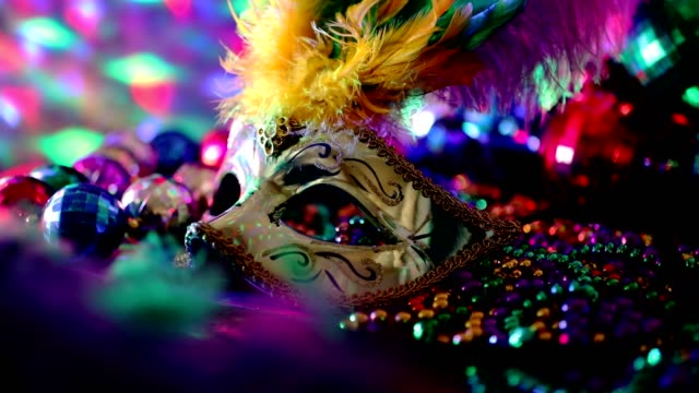 Mardi Gras or Rio Carnival mask and colorful carnival decorations.  Scene includes: gold feathered and sequined mask, colored party lights, and beads.  Objects lie on wooden table. Lockdown.  No people.