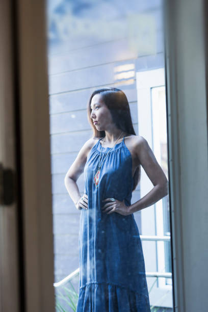 Young Asian woman outside building, hands on hips A serious young Korean woman in her 20s wearing a sleeveless blue dress, standing outdoors outside a building, hands on her hips. looking out front door stock pictures, royalty-free photos & images