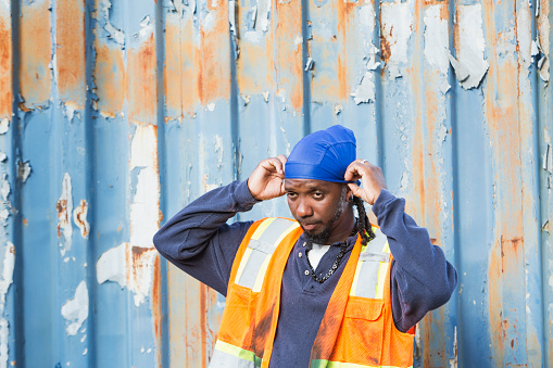 A manual worker standing in front of a rusty truck trailer wearing a reflective vest. The mature black man is putting on a do rag.