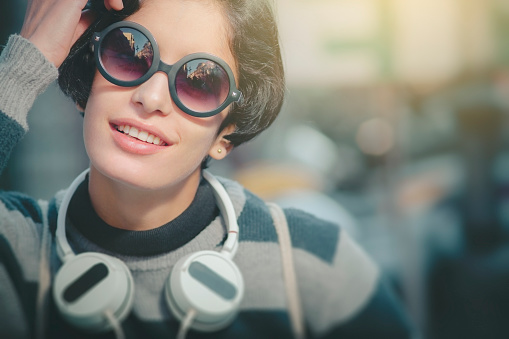 Outdoor image beautiful Asian young woman looking at camera with toothy smile. She is wearing black round sunglasses and hanging headphones in her neck. One person, waist up and selective focus with copy space.