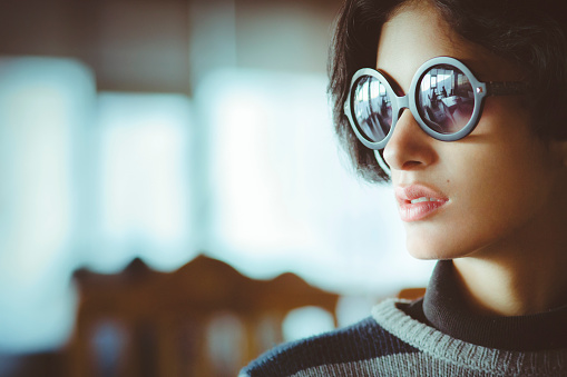 Indoor image of beautiful serene Asian young woman sitting in restaurant and looking at view. She is wearing black round sunglasses. One person, headshot and selective focus with copy space.