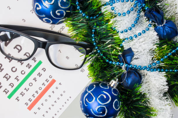 Ophthalmology or optometry Christmas and New Year with decorations. Eyeglasses and table for measure visual acuity lying near artificial snow with glitter, toys and blue balls on Christmas tree