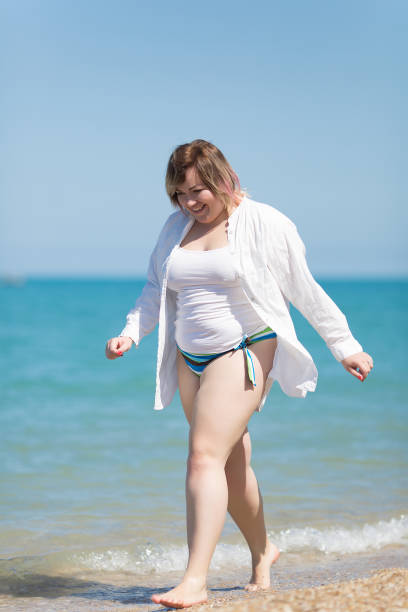 Overweight woman walking along sea. Barefoot fat girl in white shirt going on beach smiling and looking down