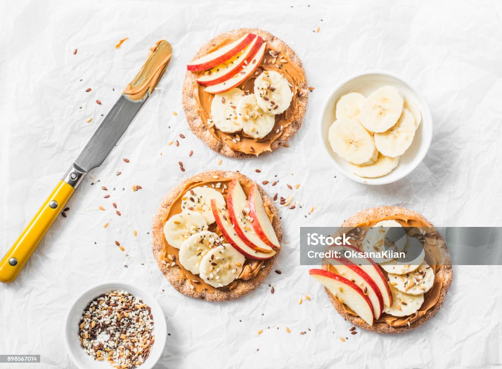 Burro di arachidi, mela, banana, cracker di semi di lino e chia brindano su uno sfondo chiaro, vista dall'alto - Foto stock royalty-free di Spuntino