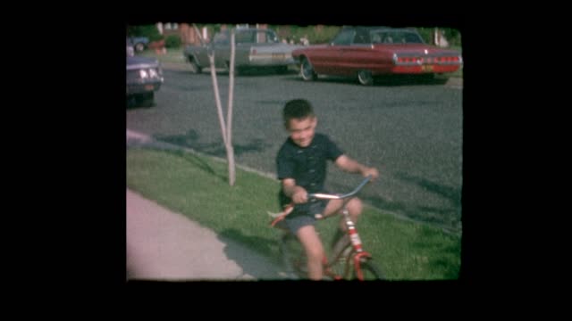 1964 little boy rides bike next to vintage cars on suburban street