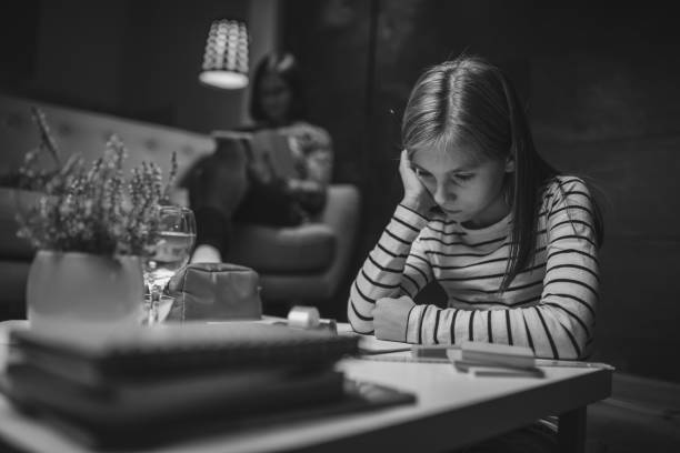 Worried girl sitting at coffee table in living room with her mother in the background and doing homework late at night