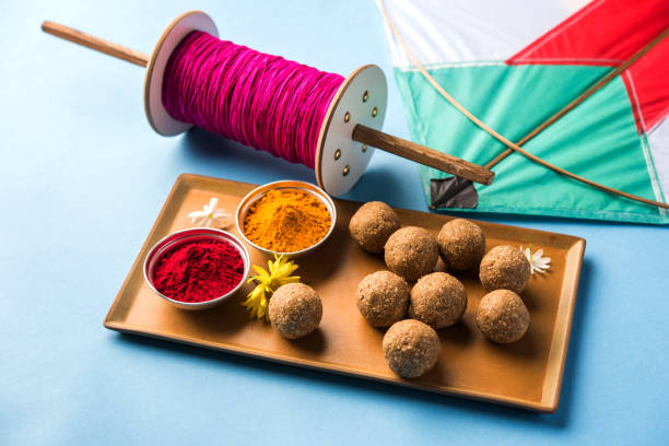happy Makar Sankranti Festival - Tilgul or Til ladoo in a bowl or plate with haldi kumkum and flowers with Fikri /Reel/Chakri /Spool with colourful thread or manjha and kite over plain background stock photo