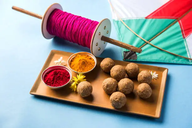 happy Makar Sankranti Festival - Tilgul or Til ladoo in a bowl or plate with haldi kumkum and flowers with Fikri /Reel/Chakri /Spool with colourful thread or manjha and kite over plain background