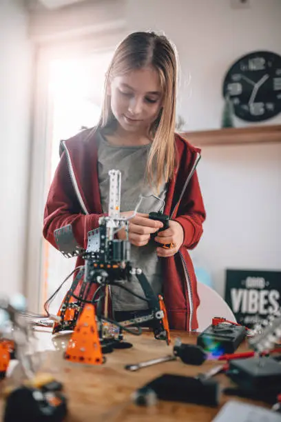 Girl wearing red hoodie building a robot on the wooden table at home as a school science project