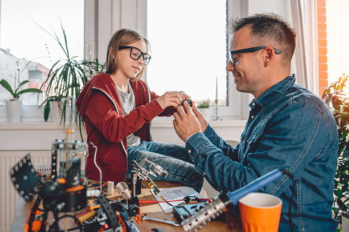 Father and daughter sitting by the wooden table and building robot at home as a school science project