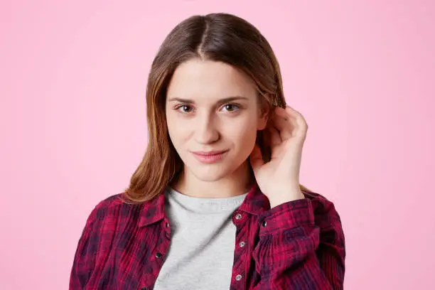 Photo of Isolated shot of adorable lovely woman has dark eyes, healthy skin, straight long hair, dressed in checkered chemise, looks confidently into camera, going to have walk in park with best friends