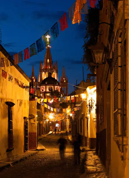 A street at night lightened by the street lights with the Parroquia of San Miguel de Allende in the background