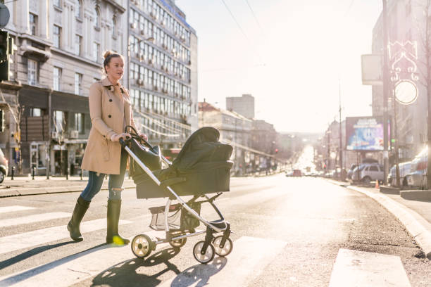 Mother pushing baby stroller on lined pedestrian crossing Side view of Young Caucasian mother enjoying with her little baby relaxing in his baby carriage. Beautiful businesswoman pushing a pram in the city street during autumn day. Brown hair mother Mother strolling with newborn, good weather for walking, healthy lifestyle. pushchair stock pictures, royalty-free photos & images