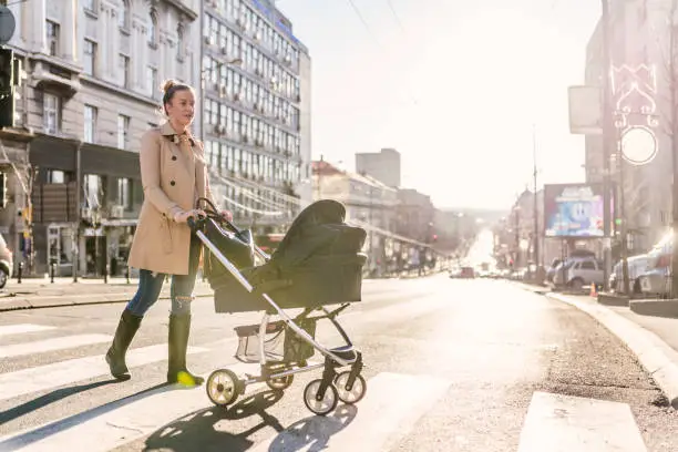 Side view of Young Caucasian mother enjoying with her little baby relaxing in his baby carriage. Beautiful businesswoman pushing a pram in the city street during autumn day. Brown hair mother Mother strolling with newborn, good weather for walking, healthy lifestyle.