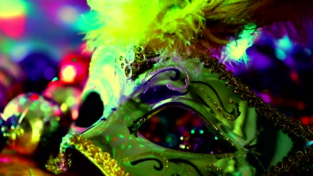 Mardi Gras or Rio Carnival mask and colorful carnival decorations.  Scene includes: gold feathered and sequined mask, colored party lights, and beads.  Objects lie on wooden table. Zoom out.  No people.