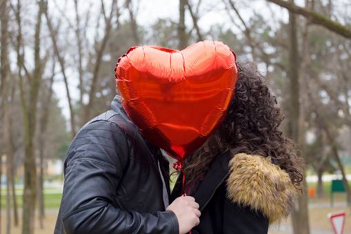 Beautiful young couple in love outdoors. Boyfriend and girlfriend hiding behind red heart shaped balloon.
