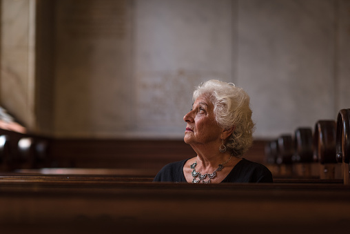 Beautiful woman on her 70s sits on a pew next to a window.