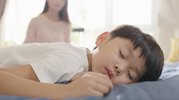 Asian boy sleeping in bed in the morning while mother sitting in background stock photo