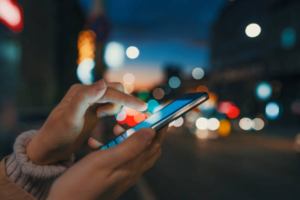 Female using her mobile phone, city skyline night light background Hand with a smart phone in a dark. Female using her mobile phone, city skyline night light background. Girl pointing finger on screen smart phone on background illumination color light in night city defocused woman stock pictures, royalty-free photos & images