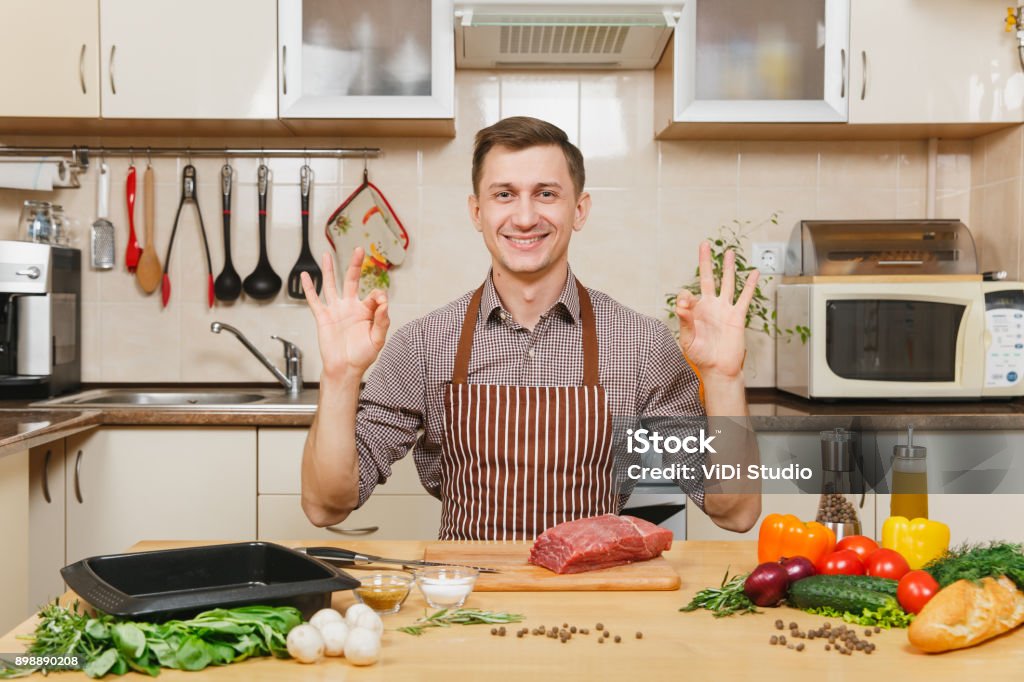 Junger Mann in Schürze mit Gemüse, am Tisch sitzen zeigt OK Geste, Kochen zu Hause vorbereiten Fleisch beteiligt von Schwein, Rind oder Lamm, leichte Küche mit Holzoberfläche, voller Phantasie Geschirr. - Lizenzfrei Beefsteaktomate Stock-Foto Junger Mann in Schürze mit Gemüse, am Tisch sitzen zeigt OK Geste, Kochen zu Hause vorbereiten Fleisch beteiligt von Schwein, Rind oder Lamm, leichte Küche mit Holzoberfläche, voller Phantasie Geschirr. - Lizenzfrei Beefsteaktomate Stock-Foto