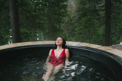 Young Caucasian woman listening to music on wireless headphones in hot tub in the forest