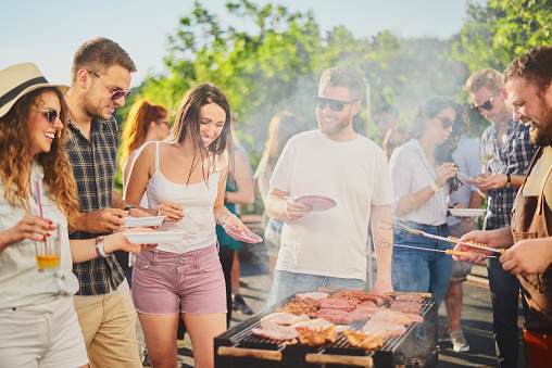 Group of people standing around grill, chatting, drinking and eating.