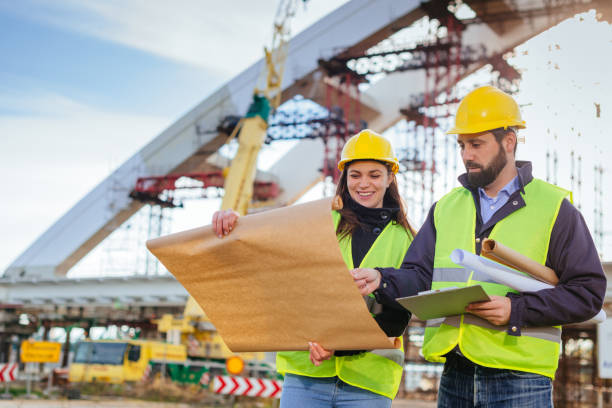 New blueprint and plans for next constuction phase Two engineers, male and female, in protective helmets and reflective clothing, standing on construction site where large arch bridge is being built. Engineers looking at blueprints, planning and discussing project development. civil engineer stock pictures, royalty-free photos & images